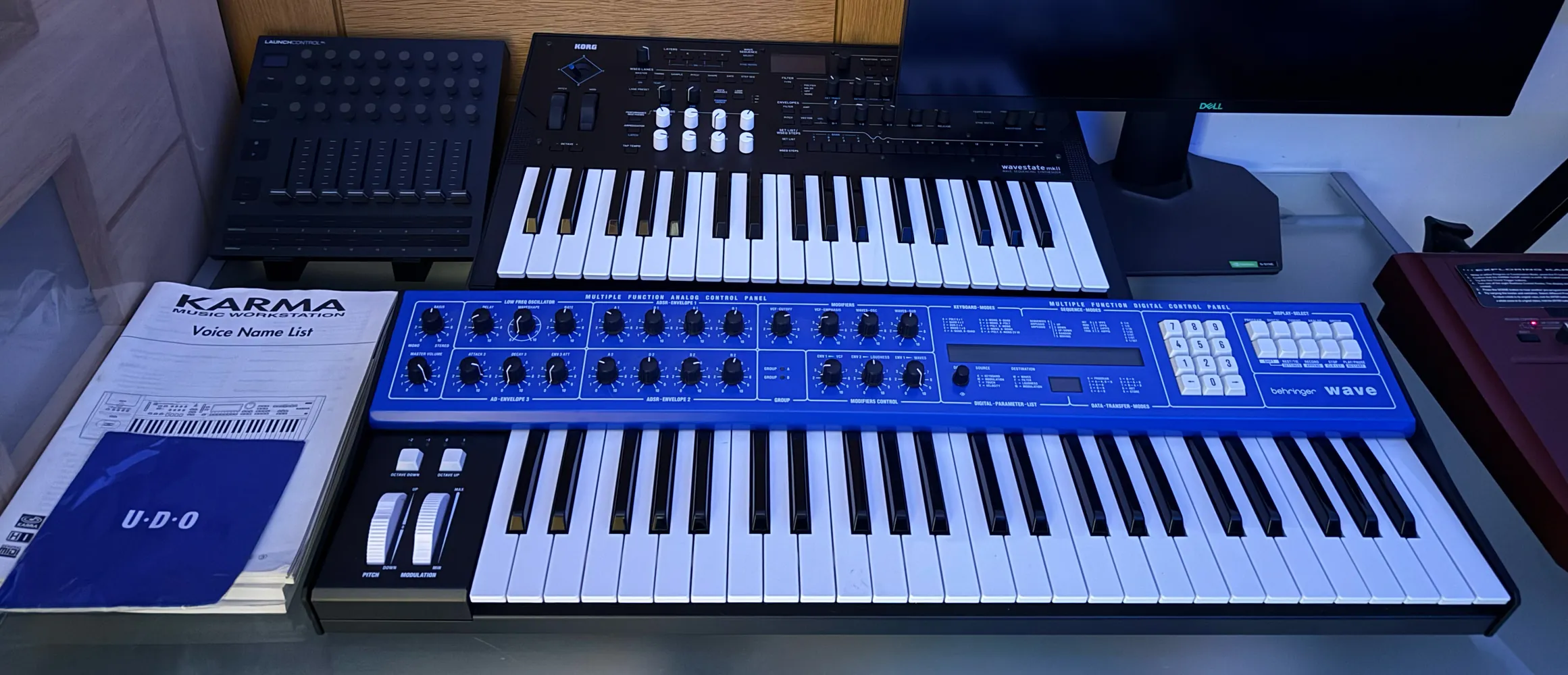 A desk with a blue synthesiser keyboard at the front, a smaller keyboard and a controller behind it and a monitor to the side.